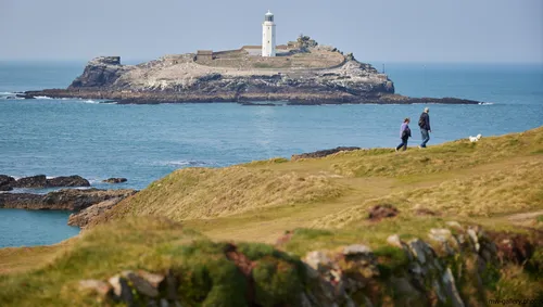 This landscape photograph captures Godrevy Lighthouse, the main subject, situated on a rocky island off the coast of Cornwall, United Kingdom. The image was taken in the early afternoon during spring, reflecting the clear visibility and gentle light typical of that season. The lighthouse, a prominent coastal landmark, stands above rugged cliffs and is surrounded by the blue waters near Sennen Cove. Nature is evident in the grassy foreground and the coastal scenery, with two people and a dog visible walking along the cliffs. The scene highlights the natural beauty of Cornwall's coastline, with Godrevy Lighthouse serving as an iconic structure along this stretch of the United Kingdom's coast.